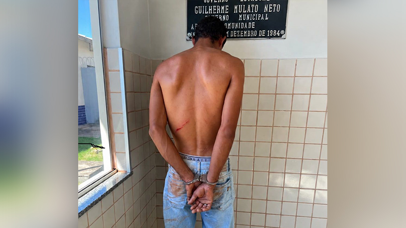 Homem detido e algemado em frente a uma parede de azulejos.