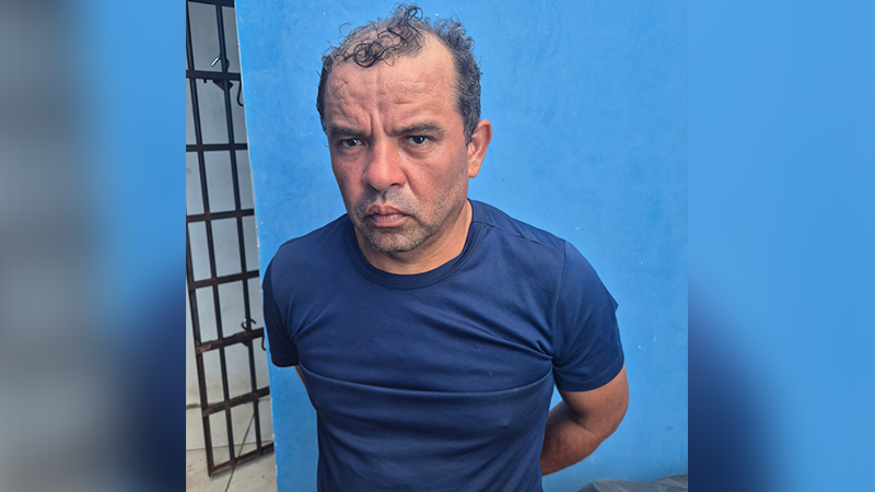 Homem de meia-idade vestindo camisa azul em frente a uma parede azul com grade ao lado.