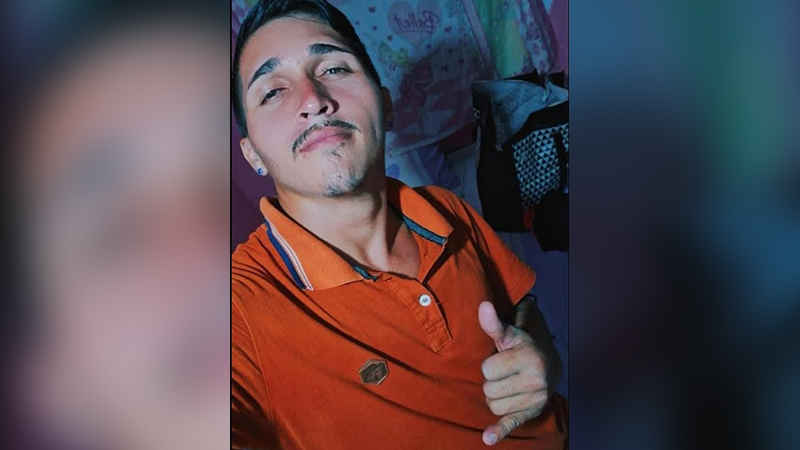 Jovem de pele clara, com bigode e cavanhaque, vestindo camisa polo laranja e fazendo gesto de positivo.
