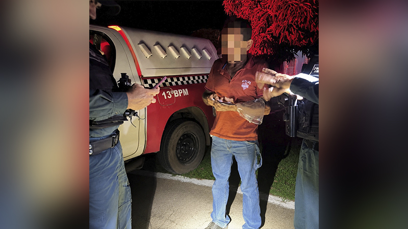 Policial militar abordando homem em frente a uma viatura do 13º BPM durante a noite.