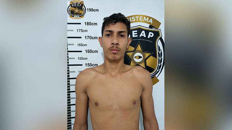 Jovem de pele clara sem camisa posando para uma foto de ficha criminal em frente a uma régua de altura com o logotipo da SEAP.
