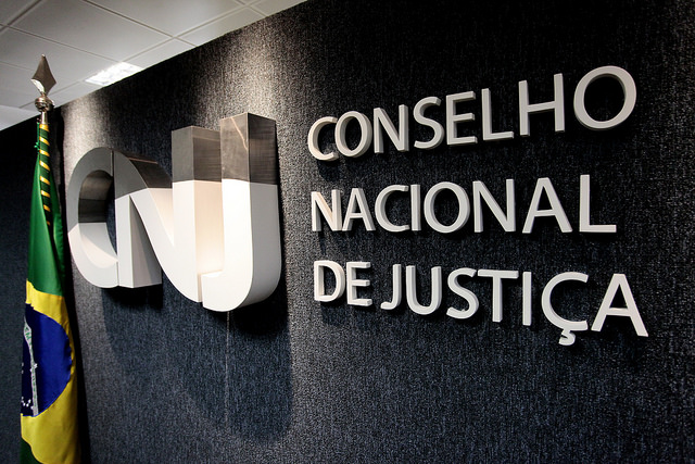 Logotipo e nome do Conselho Nacional de Justiça (CNJ) fixados em uma parede escura com uma bandeira do Brasil ao lado.