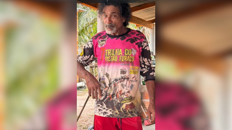 Homem de meia-idade com cabelos cacheados, barba grisalha e camiseta rosa e preta de evento de motociclismo.