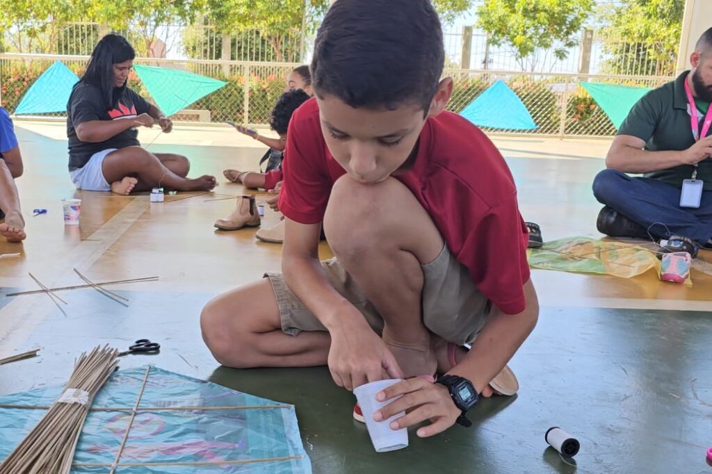 Menino de camisa vermelha sentado no chão montando uma pipa com um copo plástico