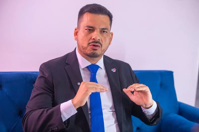 Homem de terno escuro e gravata azul gesticulando sentado em um sofá azul