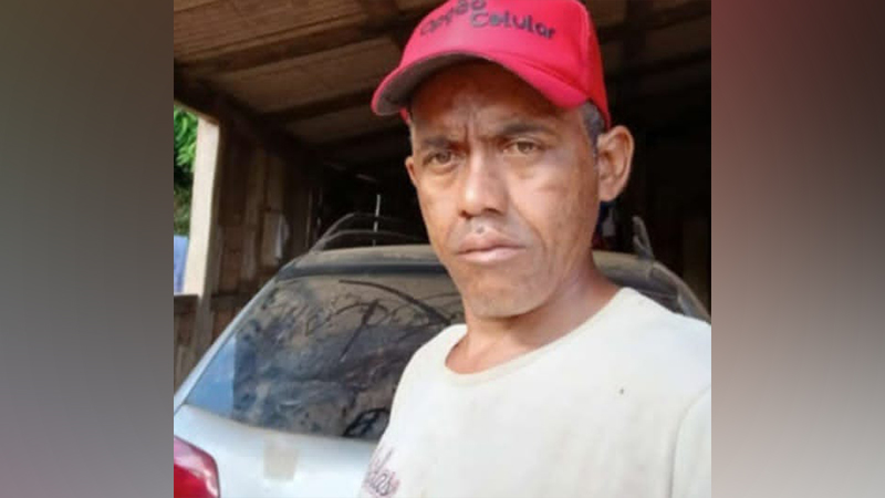 Homem de meia-idade vestindo camiseta bege e boné vermelho com a escrita 'Apoio Celular' em uma área coberta.