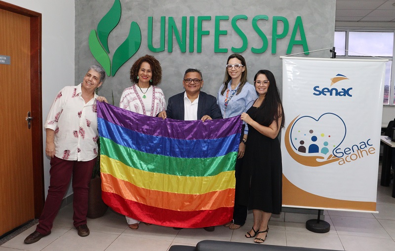 Grupo de cinco pessoas segurando uma bandeira do orgulho LGBTQIA+ em frente ao logotipo da UNIFESSPA.
