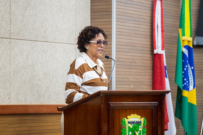 Mulher negra de cabelos cacheados curtos, vestindo blusa listrada, discursando em um púlpito de madeira com o brasão de Marabá.
