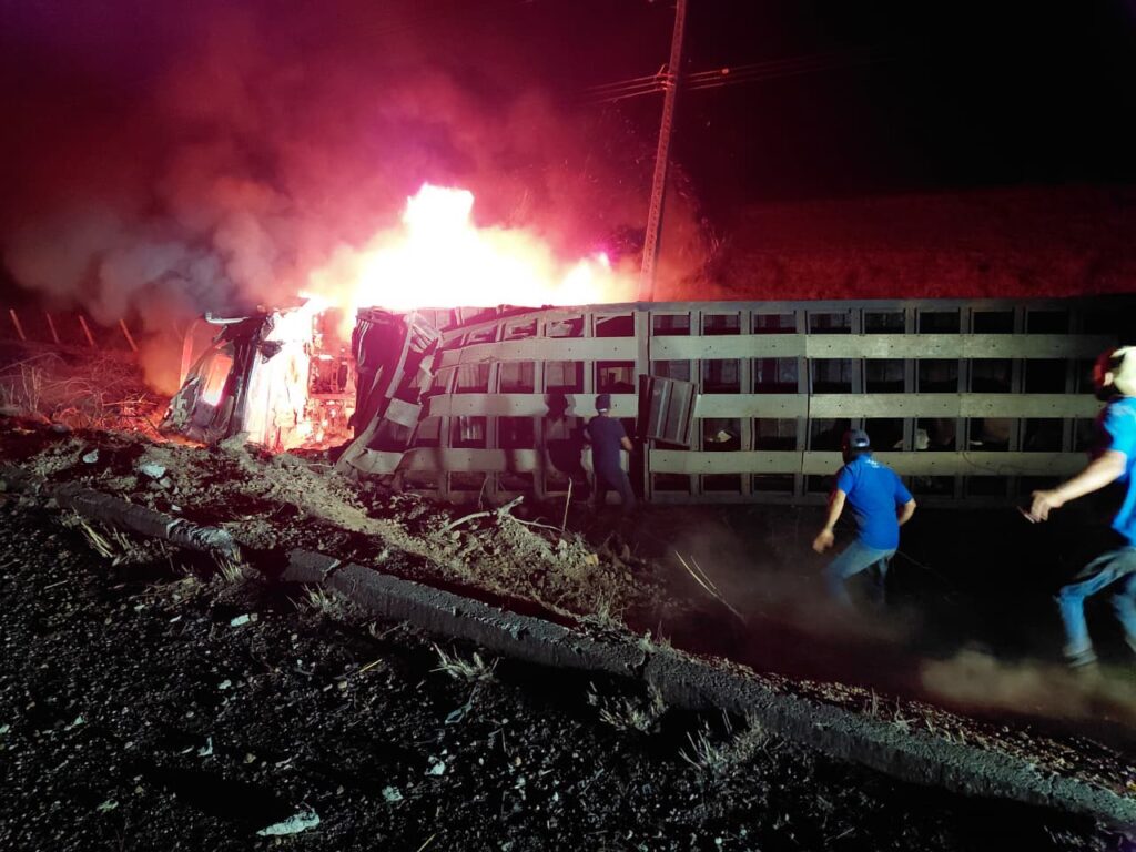 Caminhão tombado e em chamas na beira de uma rodovia durante a noite.