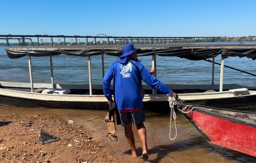 Homem de camisa azul e chapéu segurando corda de barco na beira de um rio com uma ponte ao fundo.