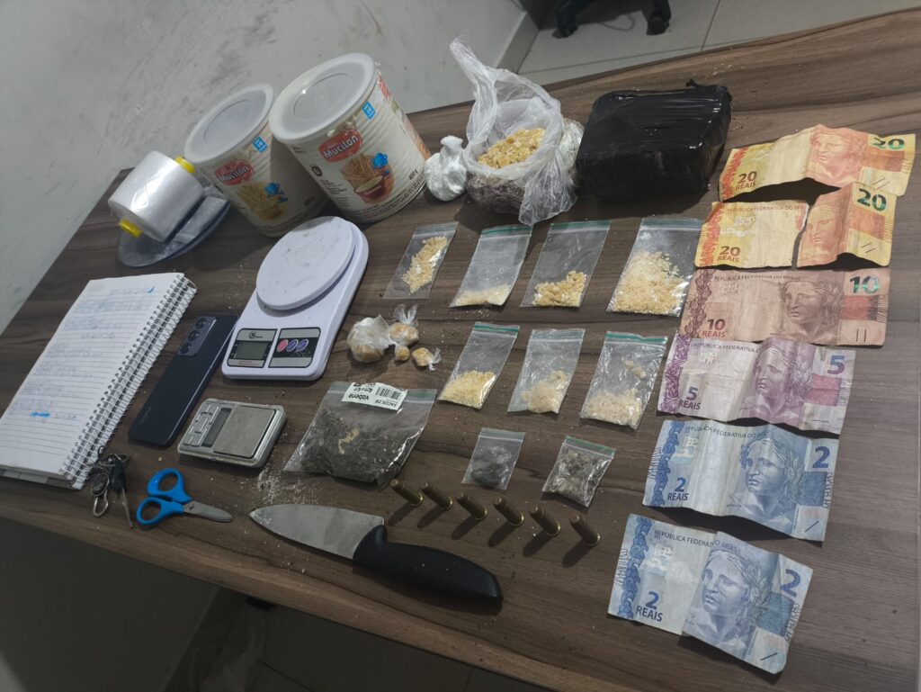 Itens apreendidos pela polícia, incluindo drogas, dinheiro, balanças de precisão e munição sobre uma mesa.