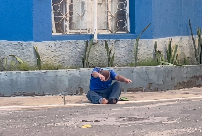 Homem sentado na calçada em frente a um prédio com parede azul.