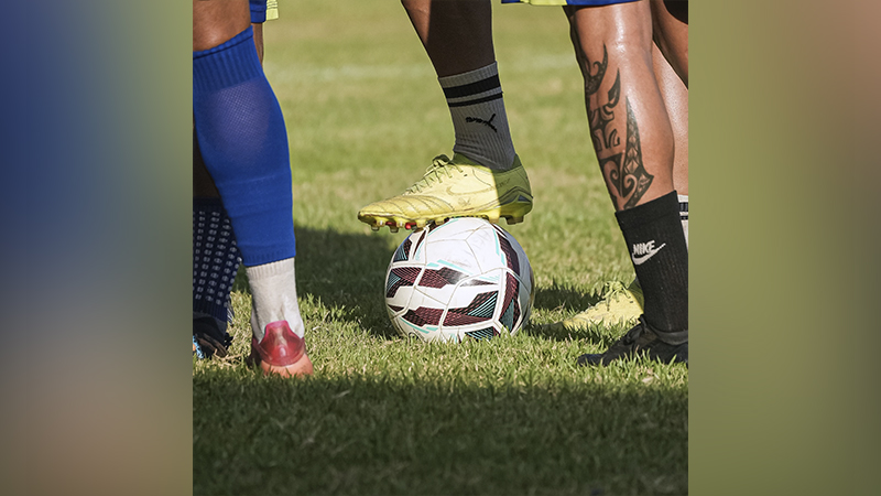 Detalhe de jogadores de futebol com os pés sobre a bola em um campo de grama.