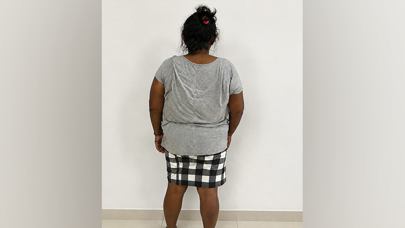 Mulher vista de costas usando blusa cinza e saia xadrez contra uma parede branca.