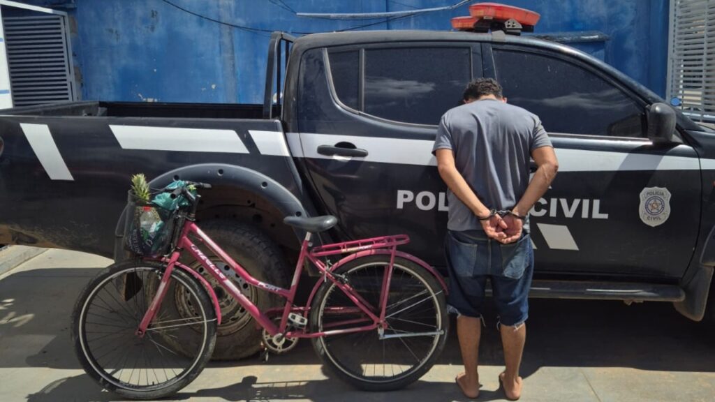 Homem algemado de costas encostado em uma viatura da polícia ao lado de uma bicicleta rosa.