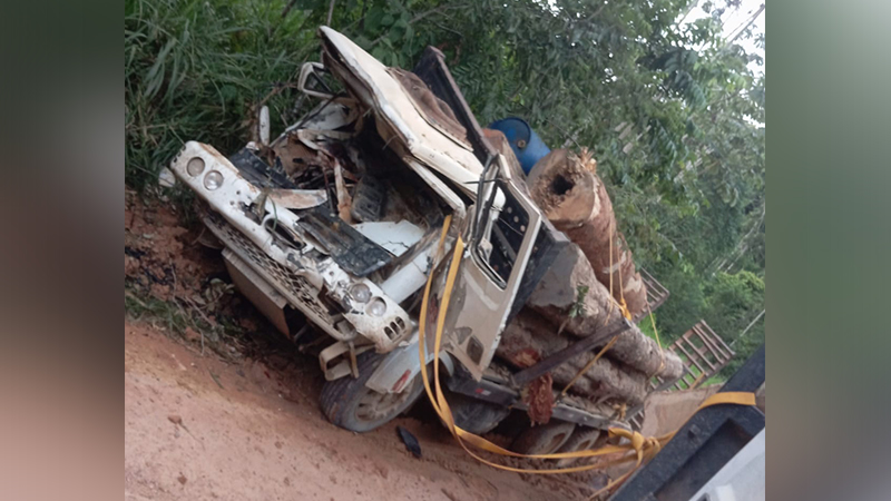 Caminhão carregado de toras de madeira tombado em estrada de terra após acidente.