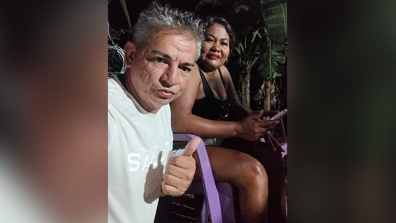 Homem de camisa branca fazendo sinal de positivo ao lado de uma mulher em um ambiente externo à noite.