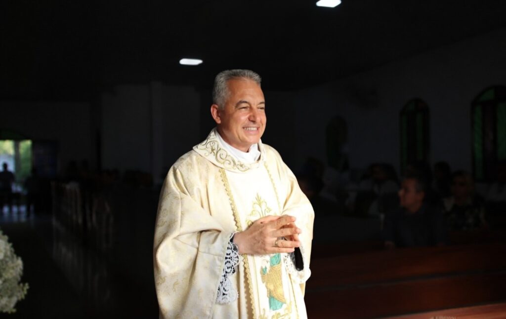 Padre sorridente vestindo paramentos litúrgicos brancos e dourados em ambiente de igreja.