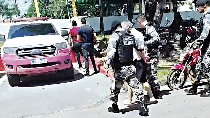Policiais militares conduzindo um homem algemado próximo a uma viatura