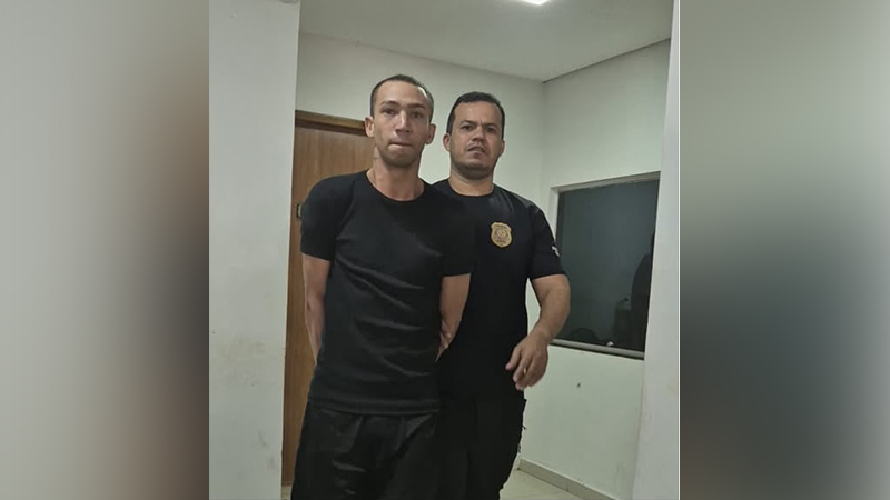 Homem algemado sendo conduzido por um agente policial em uma delegacia.