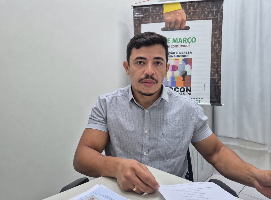 Homem de camisa cinza sentado em escritório à frente de um banner do Procon.