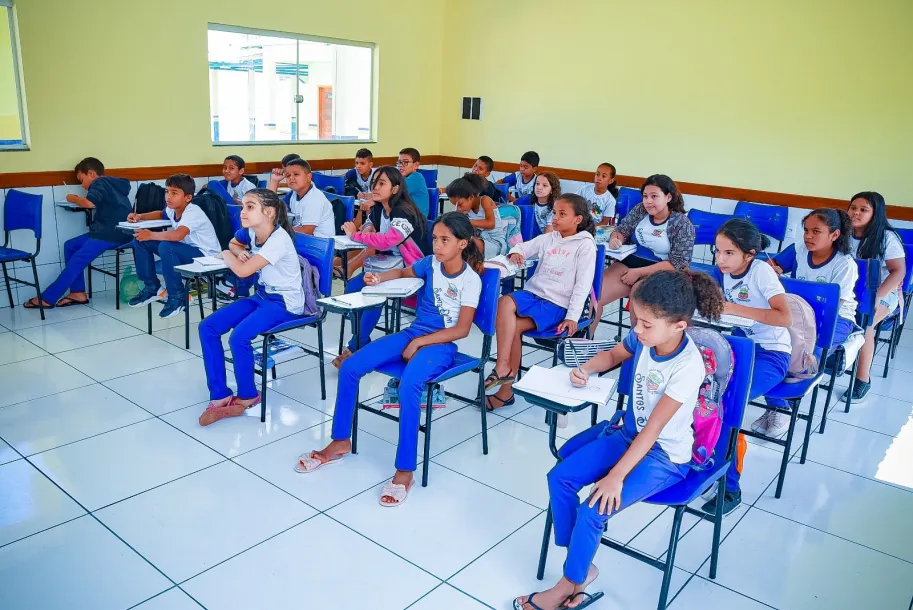 Alunos sentados em carteiras escolares em uma sala de aula iluminada.