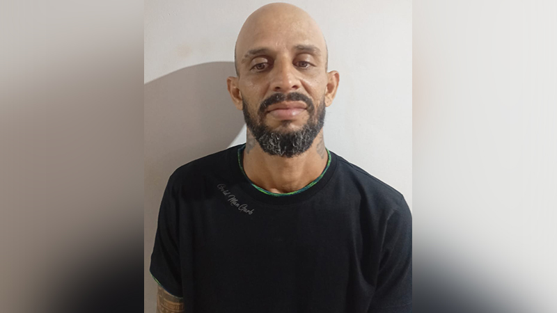 Retrato de um homem careca com barba e camiseta preta posando para foto contra uma parede branca.