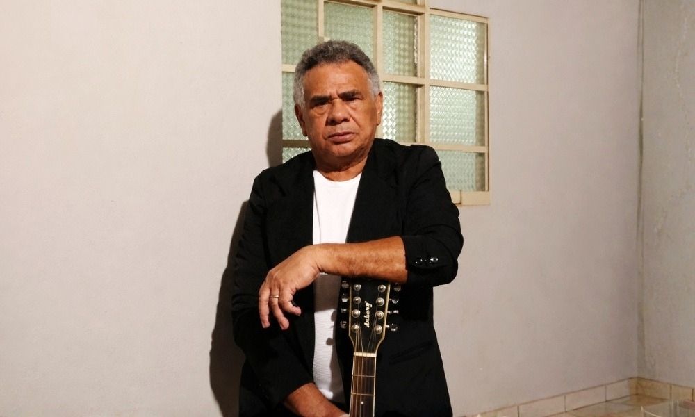 Homem de meia-idade com cabelos grisalhos vestindo blazer preto sobre camiseta branca, segurando o braço de um violão.