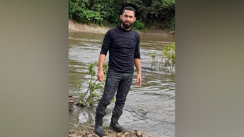 Homem com barba e camisa preta em pé na margem de um rio com vegetação ao fundo.