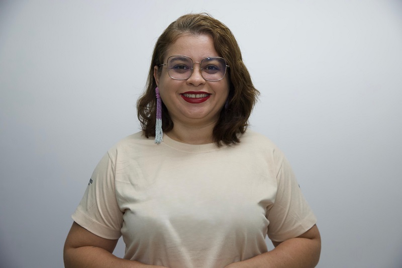 Mulher de pele clara, cabelos castanhos curtos e ondulados, usando óculos e brincos longos, sorrindo contra um fundo cinza claro.