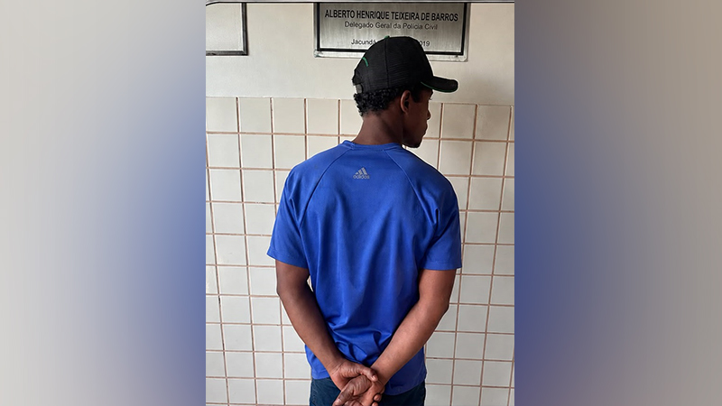 Homem de costas vestindo camiseta azul e boné, com as mãos algemadas.