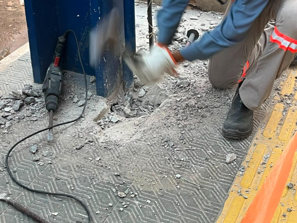 Trabalhador usando um martelo manual para quebrar o piso de concreto perto de uma coluna azul.