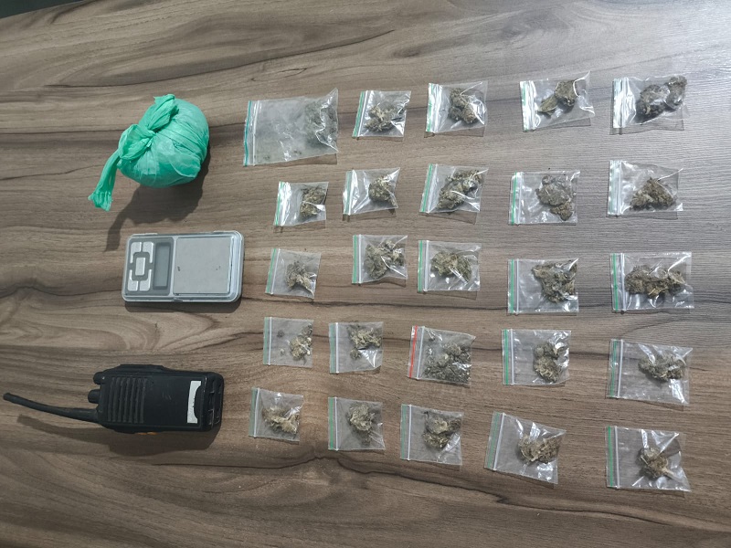 Drogas ilícitas apreendidas, incluindo pequenas embalagens de substância vegetal, uma balança de precisão e um rádio comunicador sobre uma mesa.