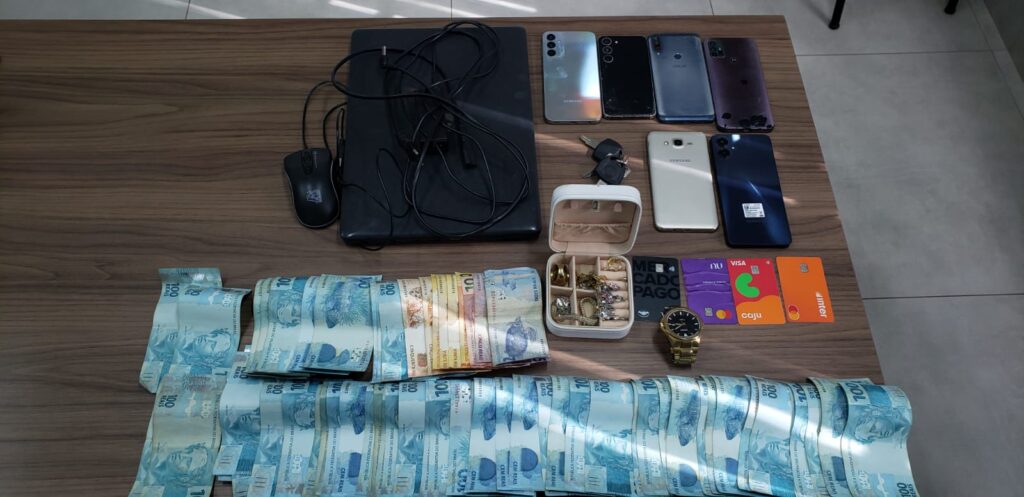 Mesa com diversos itens apreendidos incluindo dinheiro em notas de cem reais, celulares, joias e cartões bancários.