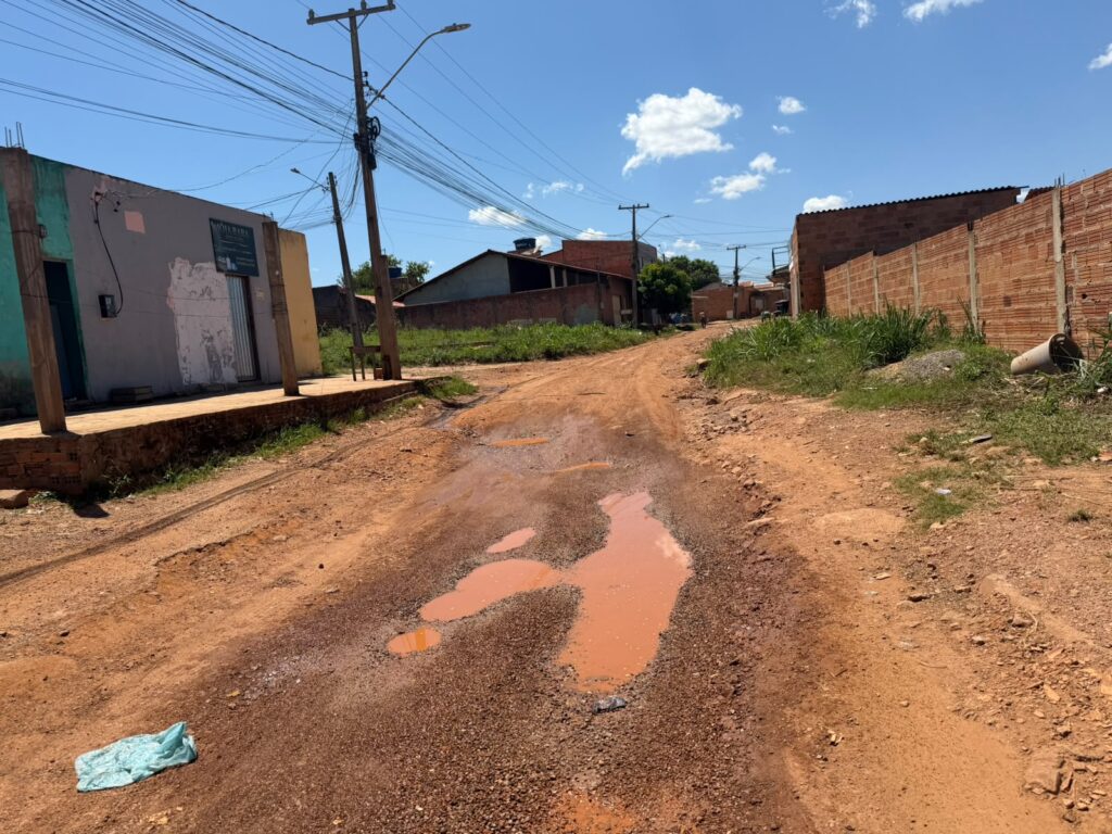 Rua de terra batida com poças de lama em bairro residencial sob céu azul.