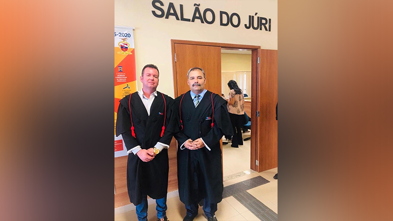 Dois homens de terno e beca de tribunal em frente a uma porta com o letreiro Salão do Júri.