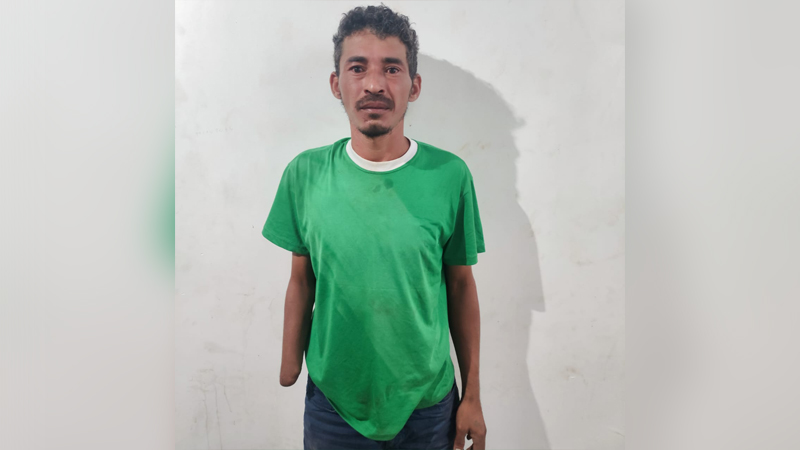 Homem de pele parda, usando camiseta verde e calça jeans, posando para uma foto contra uma parede branca.