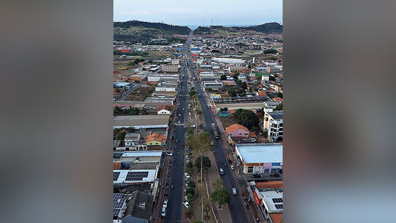 Vista aérea de uma avenida urbana em uma cidade pequena cercada por morros ao fundo.