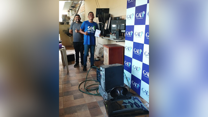 Dois homens sorridentes em um ambiente com equipamentos de informática e um banner da GAD