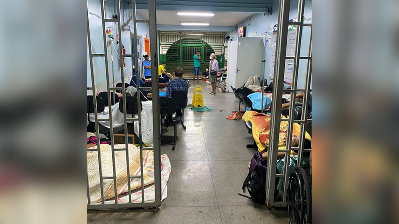 Pessoas aguardando atendimento em uma sala de espera de hospital lotada, algumas deitadas em cadeiras e no chão.