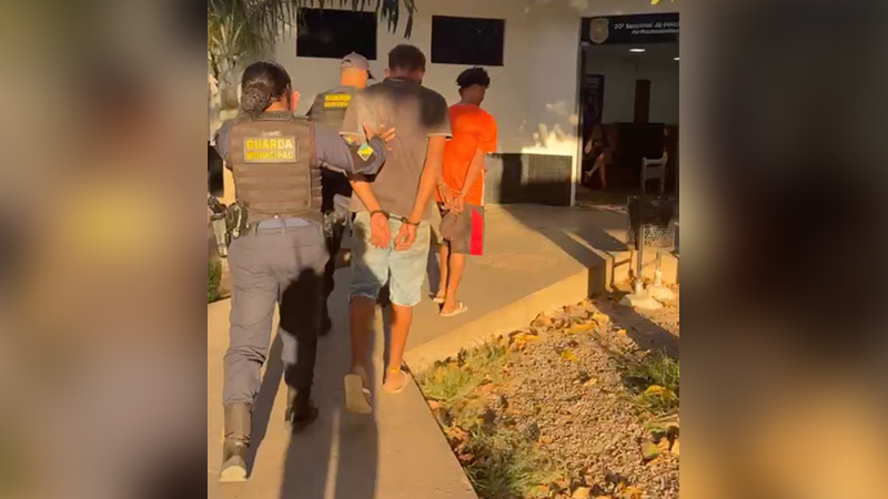 Agentes da Guarda Municipal conduzindo dois homens algemados em frente a uma delegacia.