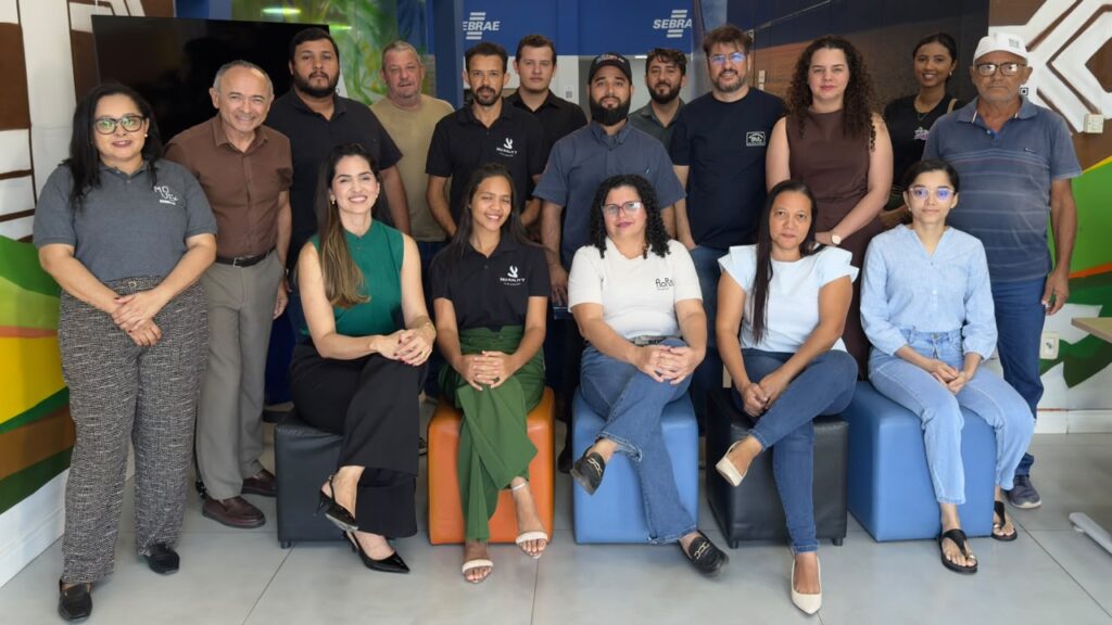 Grupo de homens e mulheres profissionais posando para foto em um ambiente corporativo com logotipos do Sebrae ao fundo.