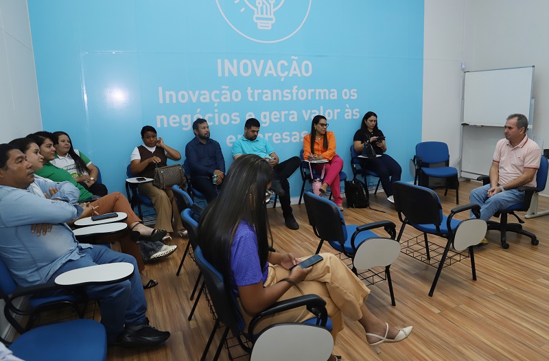 Grupo de pessoas sentadas em cadeiras universitárias durante uma reunião ou palestra em uma sala com parede azul escrita 'Inovação'.