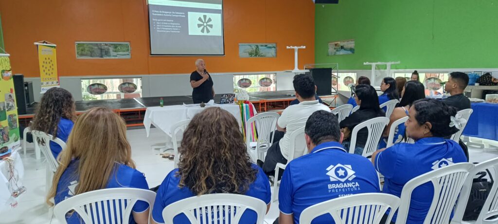 Uma palestrante em pé em um palco apresenta uma palestra para uma plateia sentada.