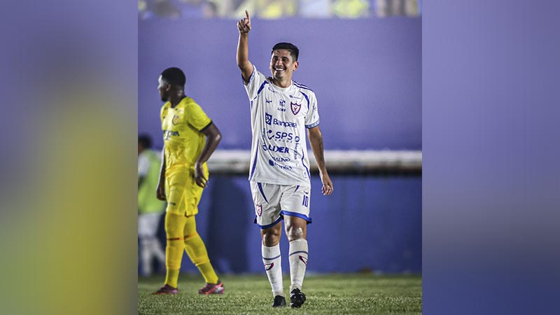 Jogador de futebol sorrindo com uniforme branco celebrando gol com o dedo apontado para o céu.