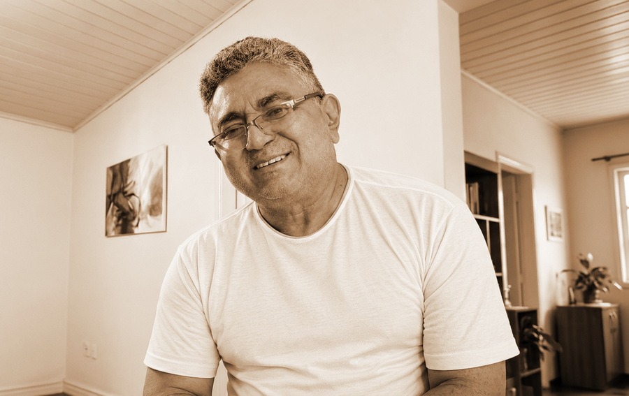 Retrato de um homem de meia-idade sorrindo, vestindo camiseta branca e óculos, com tom sépia.