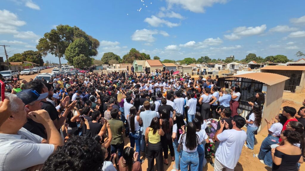 Multidão reunida ao ar livre sob céu azul em um cemitério para uma cerimônia de despedida