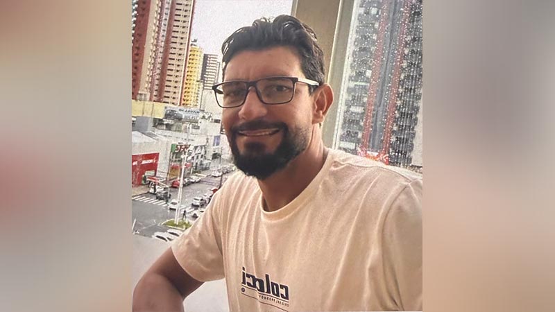 Homem de barba e óculos sorrindo em frente a uma paisagem urbana.