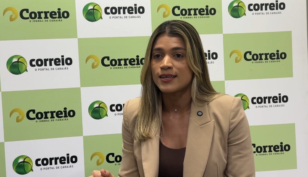 Mulher jovem com cabelos longos e castanhos em uma entrevista com fundo de logomarcas do Correio do Carajás