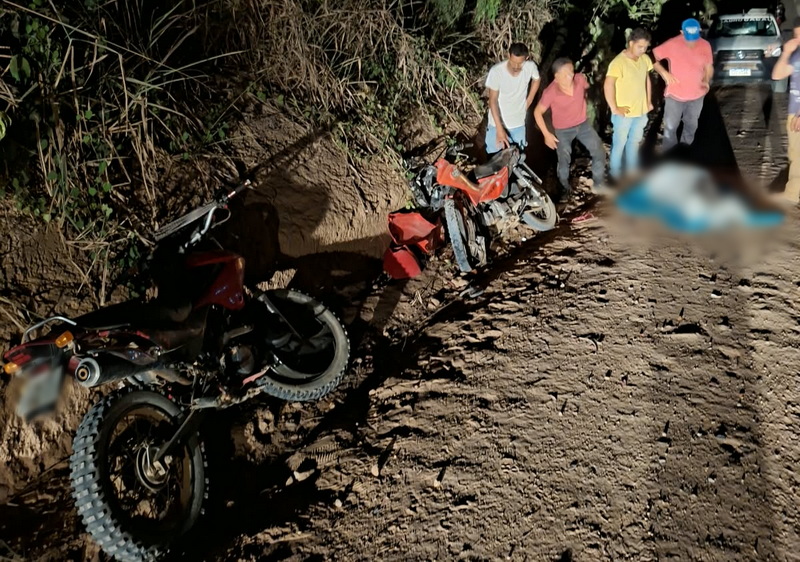 Pessoas observando motocicletas caídas na beira de uma estrada de terra à noite.